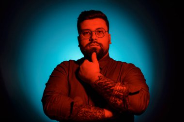 Kollarında dövmeleri ve gözlerinde gözlüğü olan sakallı bir adam, sahne ışığı etkisiyle mavi bir arkaplan önünde poz veriyor. Çenesini sol eliyle tutuyor ve kameraya bakıyor.