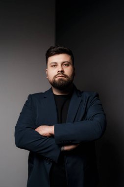 Koyu mavi takım elbiseli ve siyah gömlekli bir adam siyah beyaz bir arka plan önünde kollarını kavuşturmuş poz veriyor.