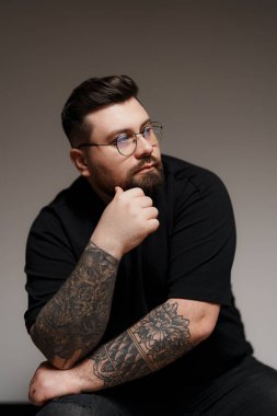 Genç, iri yarı, siyah elbiseli ve kolları dövmeli gözlüklü bir erkek model. Stüdyoda gri arka planda oturuyor.