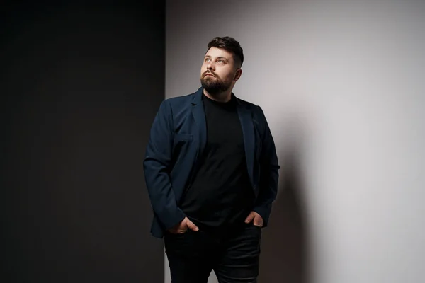Pensive young bearded male model in elegant jacket and black t-shirt standing in studio with hands in pockets and looking away thoughtfully