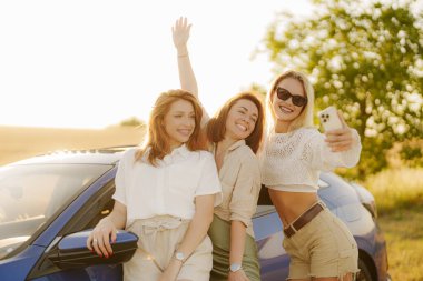 Keyifli genç kadınlar günlük giysiler içinde gülümseyip kameraya bakarken arabanın yanında durup tatildeyken cep telefonlarından selfie çekiyorlar.