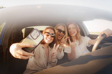 Şık gözlüklü mutlu genç kadınlar arabada oturur, parmaklarıyla gülümserken akıllı telefondan selfie çeker ve ekrana bakarlar.