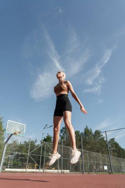 Kırmızı spor sutyenli ve siyah şortlu bir kadının mavi gökyüzü ve arka planda yeşil ağaçlarla kırmızı bir basketbol sahasında zıpladığı düşük açılı bir çekim.