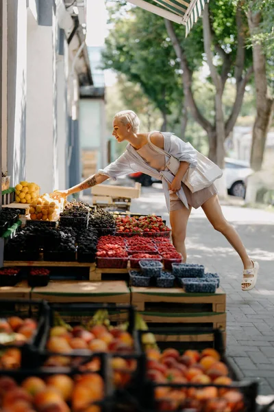 A woman reaching for some peach at a fruit stand on the street. Variety of fruits such as grapes, strawberries, blueberries and peaches. The woman interest in the fresh fruit