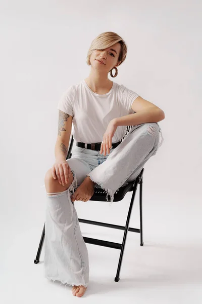 stock image Woman sitting on a black chair with her hands on her knees. She is wearing a white t-shirt, ripped jeans. She has tattoos on her arms and is posing in a relaxed manner