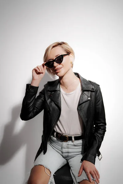 A fashionable woman poses confidently for the camera. She wears a black leather jacket, sunglasses, and ripped jeans. Minimalist and white background contrasts with her dark clothing