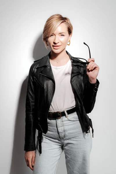 Stylish and confident young woman posing in front of a white wall wearing a black leather jacket. She is holding a pair of sunglasses in one hand and looking at the camera with a confident expression