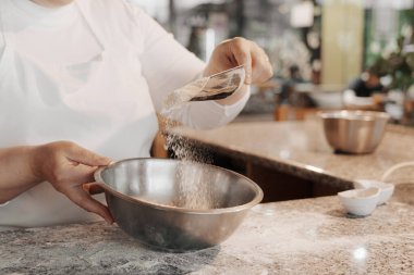 Beyaz önlüklü isimsiz kadın aşçı mutfaktaki hamuru hazırlarken metal kaseye un döküyor.