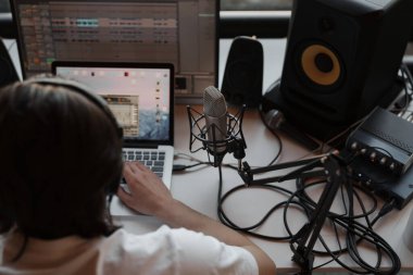 Modern stüdyoda podcast yaparken dizüstü bilgisayarda yazan beyaz tişörtlü ve kulaklıklı odaklanmış erkek müzisyenin yan görüntüsü