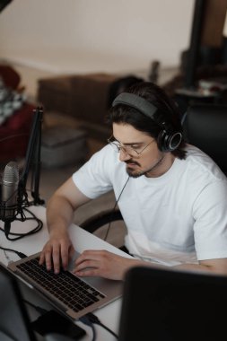 Kulaklıklı, sakallı genç bir müzisyen iş günü kayıt stüdyosunda dizüstü bilgisayarında müzik besteliyor.