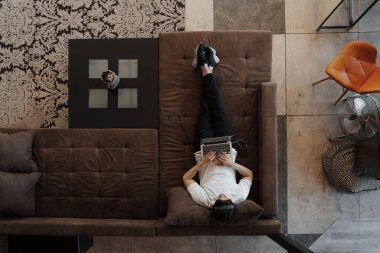 Beyaz tişörtlü, kulaklıklı, kahverengi koltukta uzanan ve dizüstü bilgisayarında müzik besteleyen isimsiz erkek müzisyenlerden oluşan üst düzey bir görüntü.