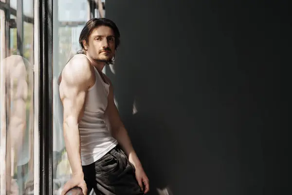 stock image Muscular man with long hair in white sleeveless t shirt and black pants standing near panoramic window and leaning on railing while looking away