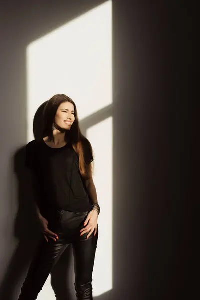 一位身穿休闲黑色服装的女性一边微笑着 一边享受着从窗户流过的自然阳光的温暖 这就产生了一种引人注目的光影相互作用 — 图库照片