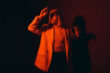 Genç bir kadın dikkat çekici kırmızı bir ceketle özgüvenle poz veriyor, silueti dramatik arka ışıklandırmayla büyütülüyor.