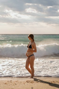 Sahilde tefekküre dalmış hamile bir kadın, altın saat ışığında güneşleniyor.