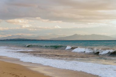 Dalgalar ve bulutlu bir gökyüzünün altında dağlık bir zemin ile geniş bir sahil.