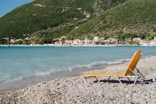 A single beach chair faces the calm sea, with a coastal village set against rolling hills in the background