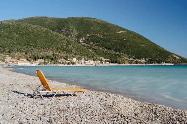 A single beach chair faces the calm sea, with a coastal village set against rolling hills in the background