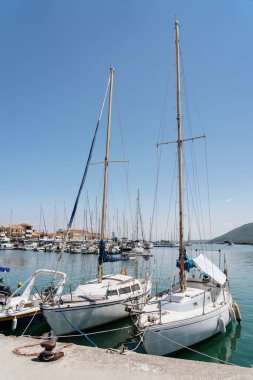 Huzurlu bir marinaya yanaşan yelkenli teknelerinin direkleri, sakin bir sahil kıyısındaki açık mavi gökyüzünü işaret ediyor.