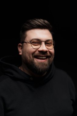 Şık gözlüklü neşeli bir adam siyah kapüşonlu bir kapüşonluyla karanlık bir arka planda mutluluk yayıyor.