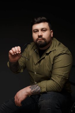 Derin düşüncelere dalmış bir adam gözlüğünü tutar, dalgın tavırlarını tamamlayan bir zeytinli gömlek.