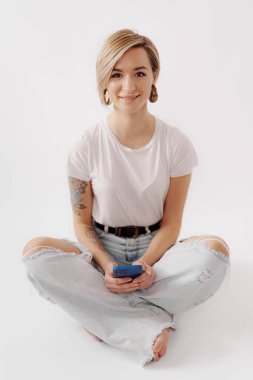 Hoş bir gülüşü olan genç bir kadın, elinde telefon tutarken bacak bacak üstüne atmış, hafif ve havadar bir şekilde.