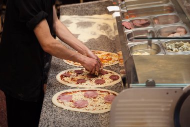 Siyah önlüklü bir şef, pizzalara titizlikle malzeme ekler. Elinde paslanmaz çelik bir malzeme tezgahı vardır.