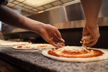 Şefin ellerinin pizza hamurunun üzerine ustalıkla peynir serptiği yakın çekim görüntüsü.
