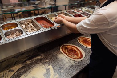Profesyonel bir mutfakta bir aşçı, pizza hamuru üzerine çeşitli malzemeler hazırlar ve arka planda çeşitli malzemeler bulunur.