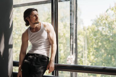 Askılı tişörtlü düşünceli bir adam, yansıtıcı bir ifadesi ve doğal bir arka planı olan güneşli bir pencereden dışarı bakar.