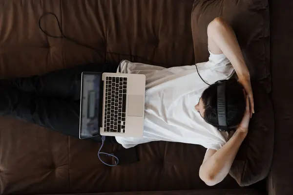 A top-down shot of a person lying down on a couch with a laptop, taking a break or contemplating while working from home