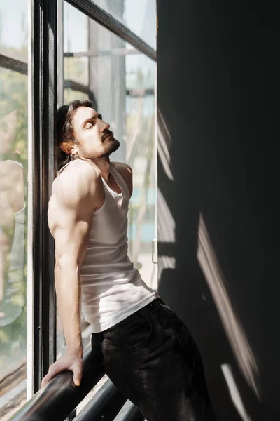 A man in a tank top leans against a window, basking in the warm sunlight, eyes closed in relaxation