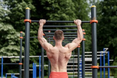 Arka planda kaslı bir sporcunun açık hava spor aletleri üzerinde barfiks çekerken güçlü sırt kasları göstermesi.