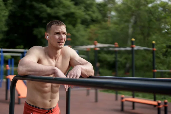 Shirtless athlete leans on parallel bars at an outdoor gym, lost in thought after a rigorous training session