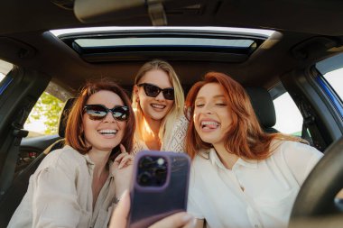 Arabada üç kadın var, mutlu bir şekilde poz verip selfie çekiyorlar..