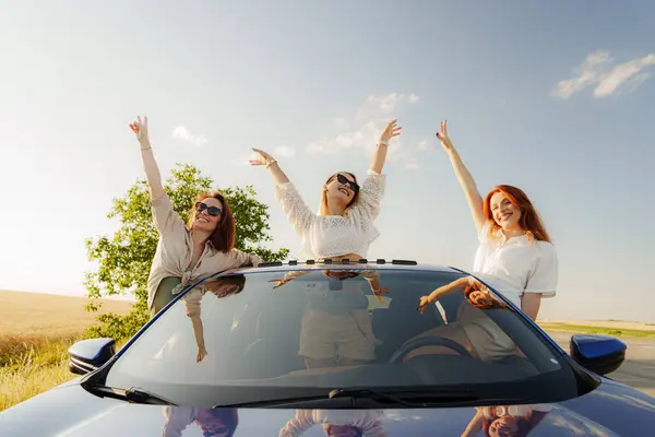 Three friends pop out of a car sunroof, arms raised in joy, embracing the freedom of a summer road trip in the countryside