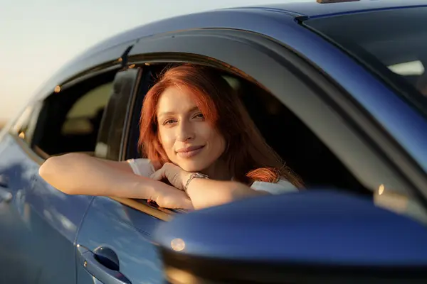 Red-haired woman resting on car window during a golden sunset, embodying calmness and travel spirit