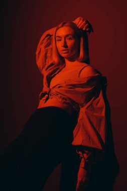 Moda mankeni kendine güvenle poz verir, gölgelerle örtülmüş ve kırmızı ışık onun pozunu vurgular.