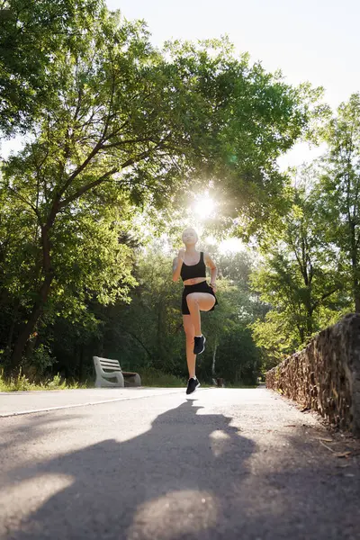 Dynamic low-angle shot of a woman jogging on a park path with morning sunlight filtering through trees