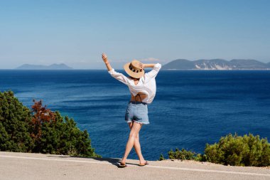 Kolları açık bir kadının kıyı manzarasından engin mavi denizin tadını çıkarırken.