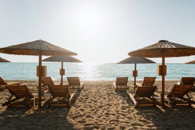 Kumlu bir sahilde, saman şemsiyelerin altında tahta güneşlikler, deniz kenarında bir gün dinlenmeye davet ediyor.