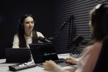 Bir kadın sunucu ve misafiri, podcast sırasında, kapalı alanda, profesyonellik ve özen göstererek, iş kıyafetleri içinde, düşünceli bir medya çatışması anı yakalıyorlar.