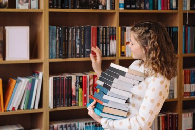 Puantiyeli bir genç kız elinde bir yığın kitapla raftan kitap seçiyor.