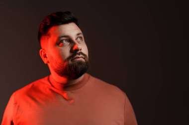 Sakallı bir adam yukarı bakar, yüzü yumuşak, kırmızı bir ışıkla vurgulanır, yansıtıcı bir ruh hali yaratır.