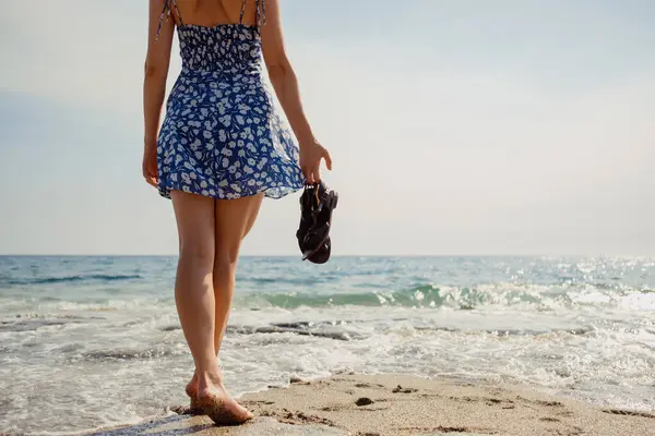 ベアフットとケアフリー 手のサンダルを持つ女性は 晴れた海岸線に沿って柔らかい砂と穏やかな波を楽しんでいます ストック写真