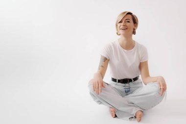 Neşeyle ışıldayan genç bir kadın dar kot pantolonla bağdaş kurmuş, zarafeti ve hayat dolu ruhu somutlaştırıyor.