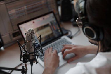 Modern stüdyoda podcast yaparken dizüstü bilgisayarda yazan beyaz tişörtlü ve kulaklıklı odaklanmış erkek müzisyenin yan görüntüsü