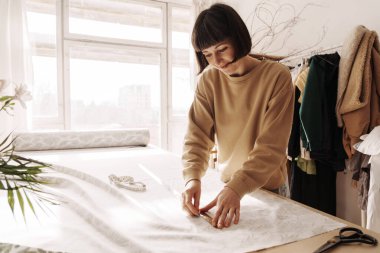 Yaratıcı kadın terzi, bitkilerle dolu iyi ışıklandırılmış bir dikiş atölyesinde masa üzerinde kumaş ölçüyor..