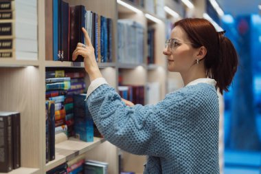 Kütüphane ortamındaki bir raftan kitap seçen odaklanmış genç bir kadın..