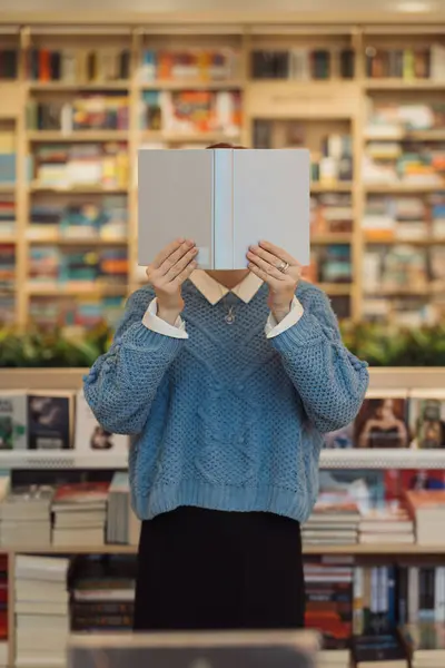 An unidentifiable person holding a book in front of face in a library.